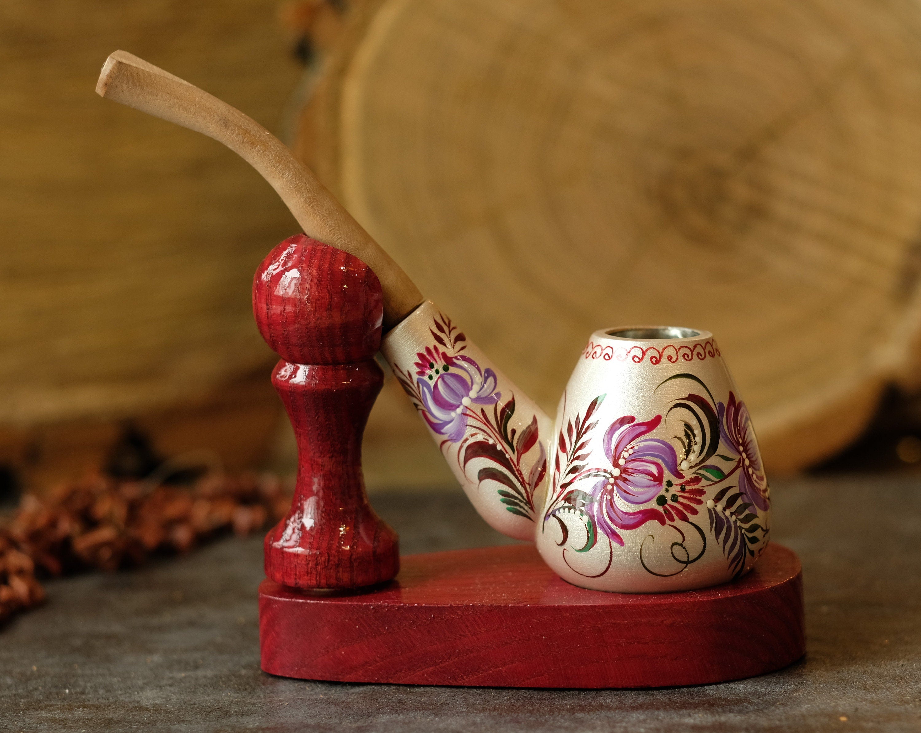 May include: A wooden smoking pipe with a light-colored bowl featuring hand-painted purple and red floral designs. The pipe rests on a dark red wooden stand with a matching red spherical support piece and a long, light-colored wooden stem.